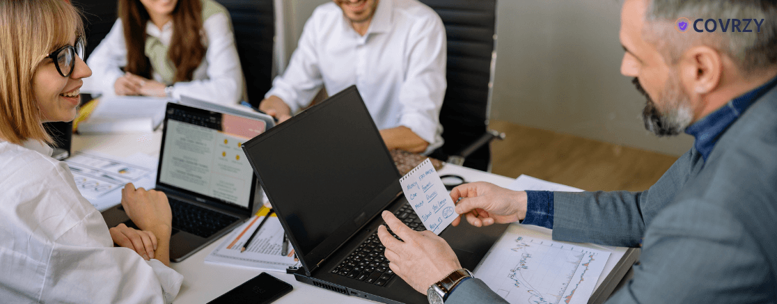 few employees sitting together with their laptops open, one of them wearing a grey suit is reading a small paper note