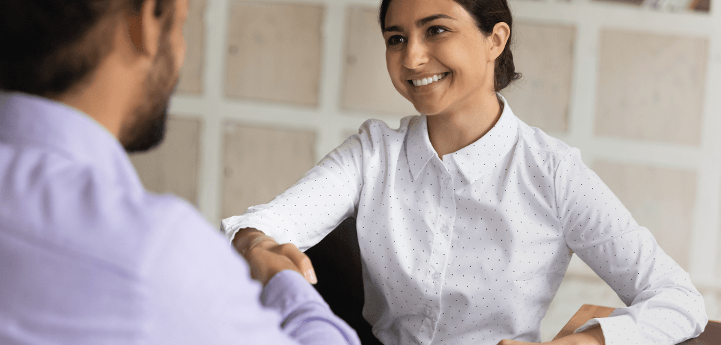 an old man in a light pink shirt shaking hands with a younger man dressed in a navy blue suit, the background is a shelf